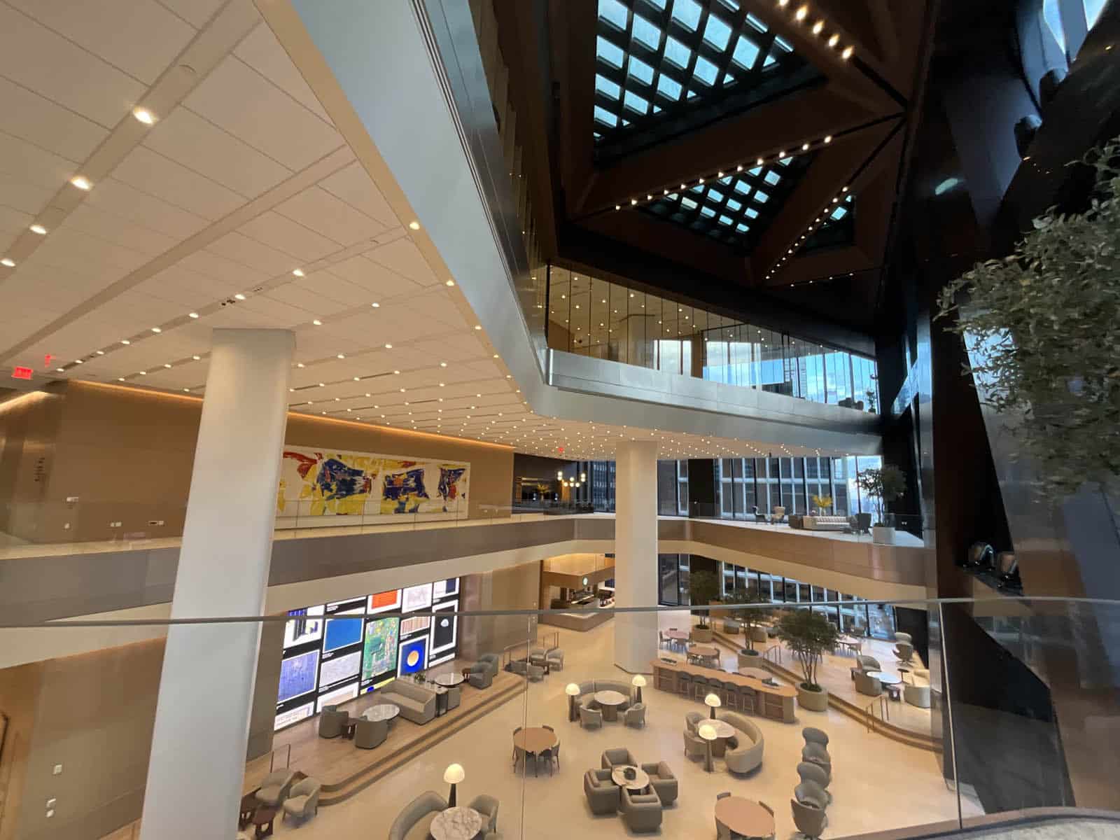 A modern, spacious hotel lobby with multiple floors, large windows, skylights, comfortable seating areas, and vibrant artwork on the walls. Warm lighting and glass railings create an open, inviting atmosphere.