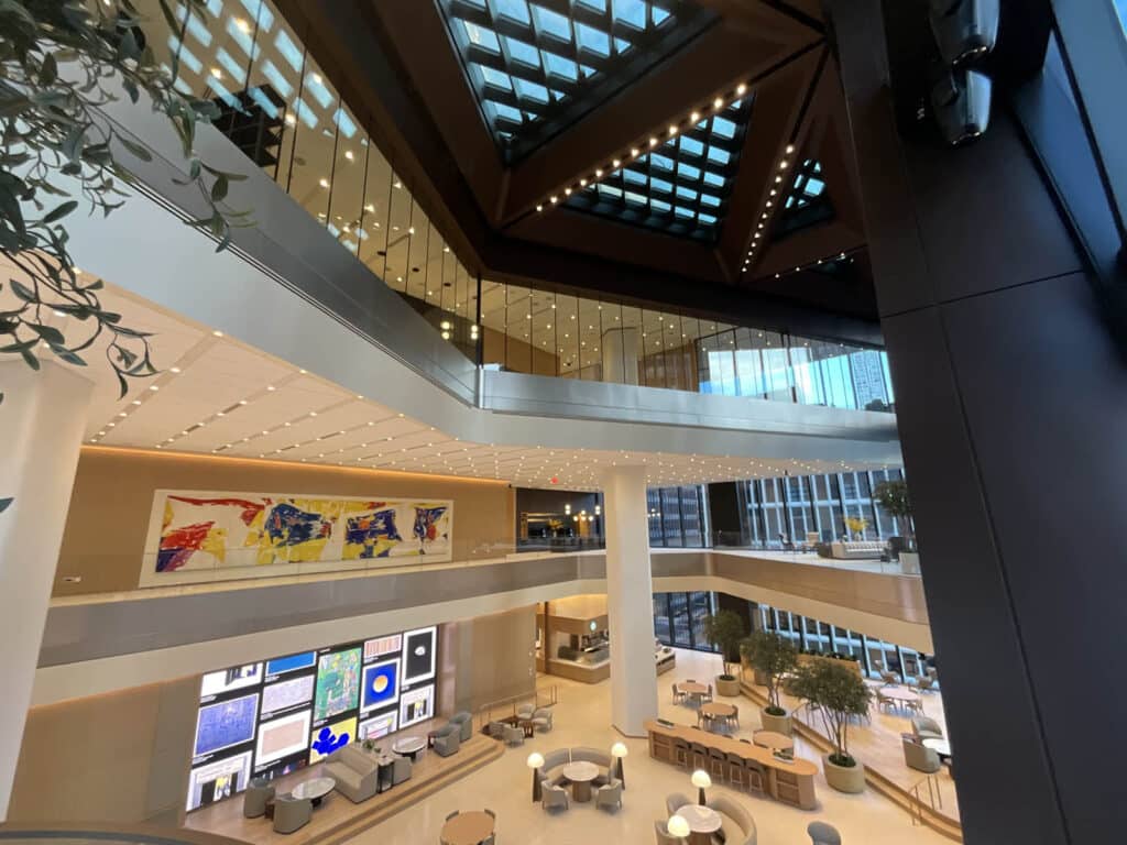 Spacious, modern atrium with large skylights, contemporary art on the walls, digital displays, round tables, and indoor trees; multiple floors are visible, with glass railings and natural lighting.