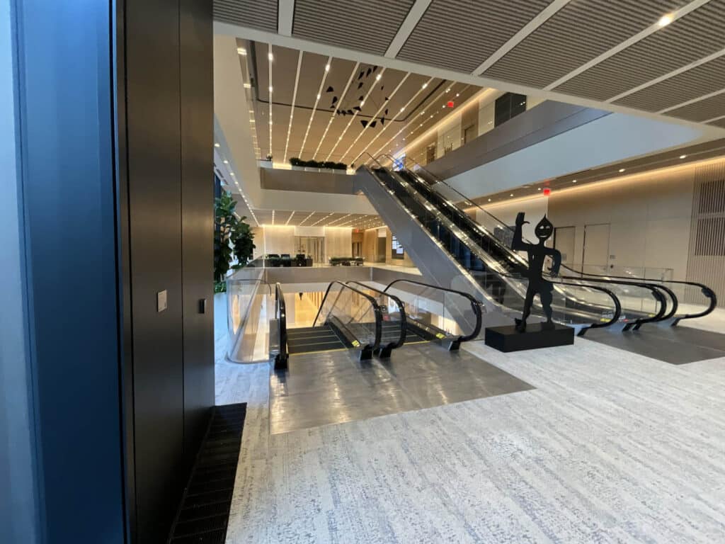 Modern office building interior with escalators going up and down, light-colored floors, a black abstract statue near the escalators, and large windows letting in natural light.