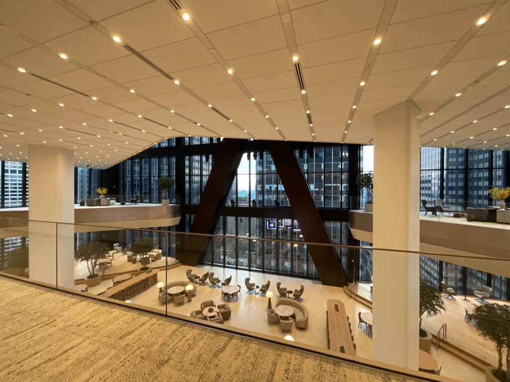 Modern office lounge with floor-to-ceiling windows, glass railing, and contemporary furniture; city skyscrapers are visible outside, and the space is brightly lit with ceiling lights.