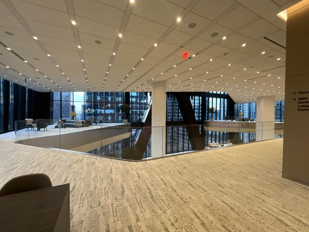 Modern office interior with large windows, light-colored flooring, glass railing, and recessed ceiling lights. The space is open, bright, and overlooks city buildings through the expansive windows.