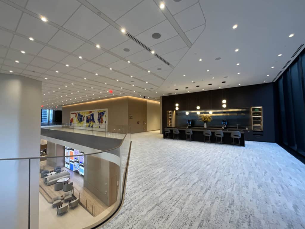 Spacious, modern office lobby with high ceilings, bright lighting, and a curved glass railing. Artwork decorates one wall, and a row of computers with pendant lights sits against a black accent wall.