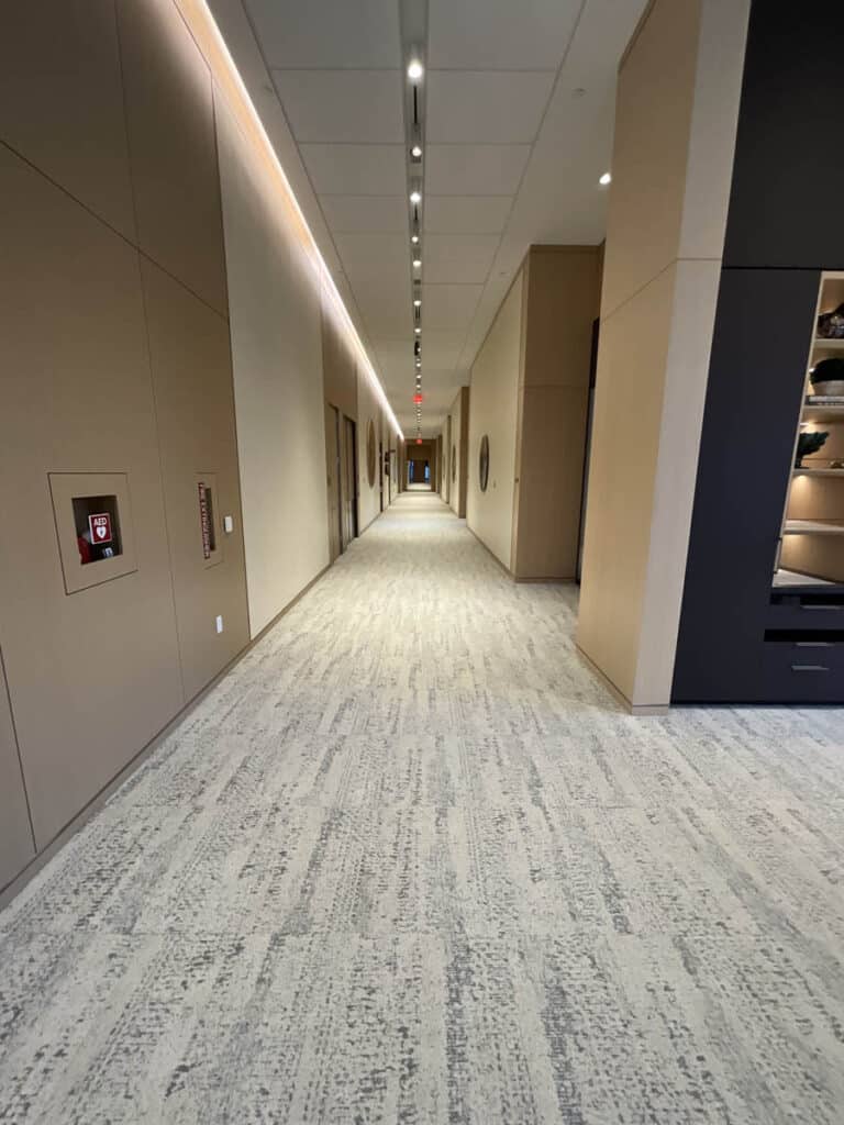 A modern, well-lit hallway with beige walls and light carpet flooring, featuring closed doors on both sides and recessed ceiling lights leading to a distant red exit sign.