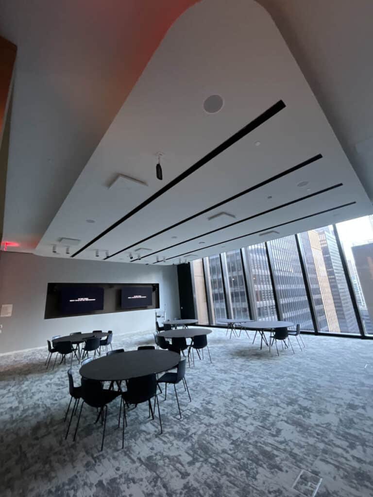 A modern conference room with round tables and black chairs, large floor-to-ceiling windows overlooking city buildings, and two wall-mounted screens at the front. The room has a patterned carpet and a high ceiling.