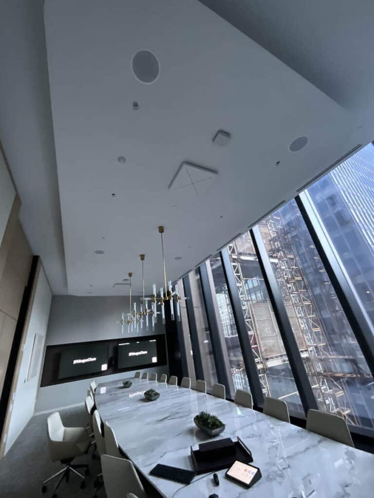 A modern conference room with a long marble table, white chairs, hanging gold light fixtures, large floor-to-ceiling windows, and a cityscape view with a construction site outside.