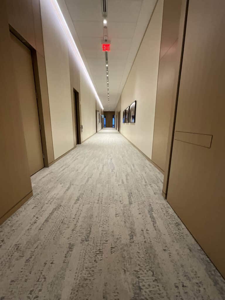 A long, well-lit hallway with beige walls and carpet, wooden doors on both sides, framed art on the right wall, and an exit sign with a red light at the far end.