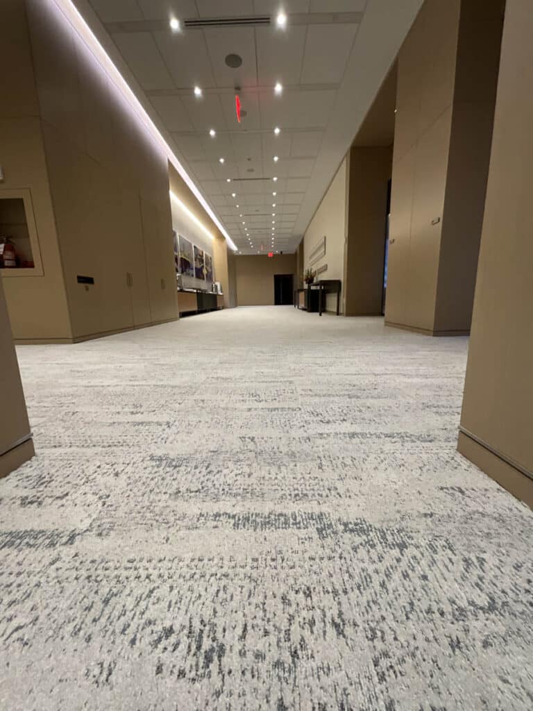 A wide, empty hallway with beige walls, a patterned light gray carpet, and a ceiling lined with recessed lights. Shelves and tables are visible along the left and right walls.
