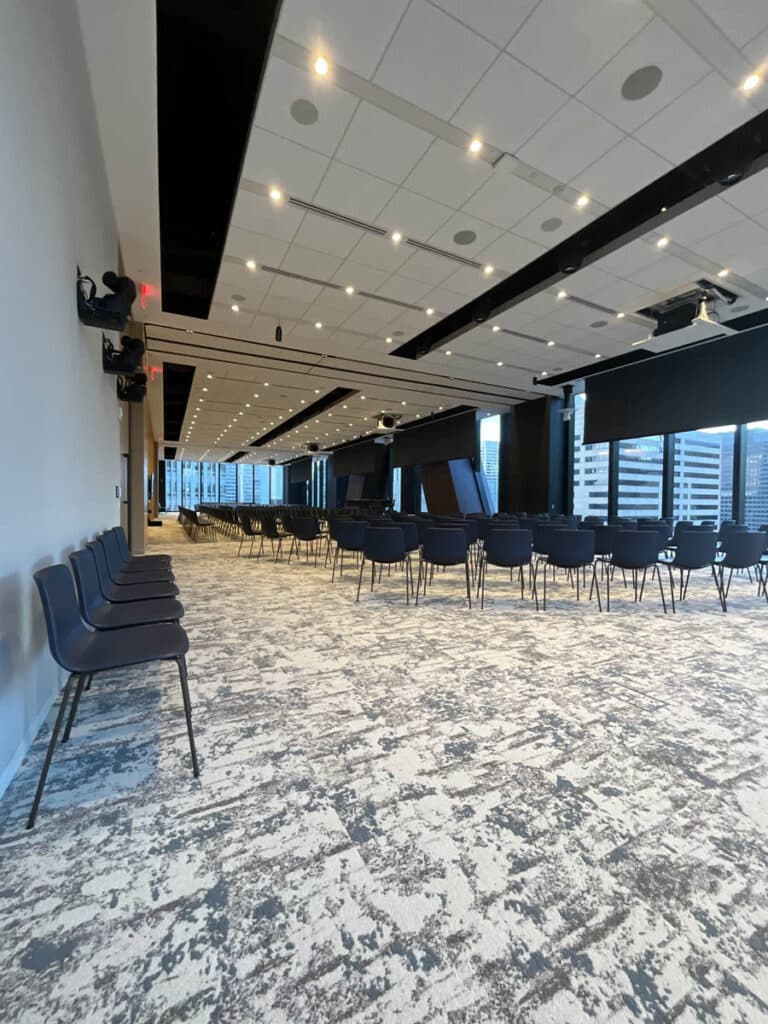 A modern conference room with rows of empty black chairs facing large windows and screens. The space features ceiling lights, patterned carpet, and city buildings visible outside.