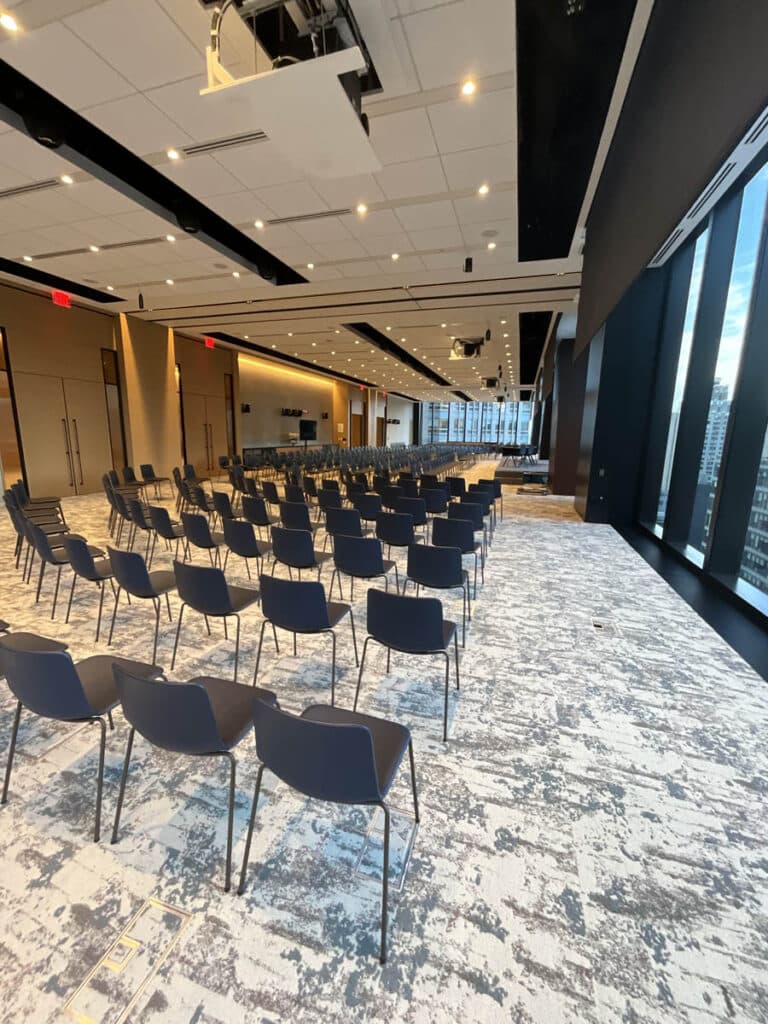 Rows of empty black chairs are arranged in a modern conference room with large windows, patterned carpet, and ceiling lights. The room overlooks a cityscape through the floor-to-ceiling windows on the right.