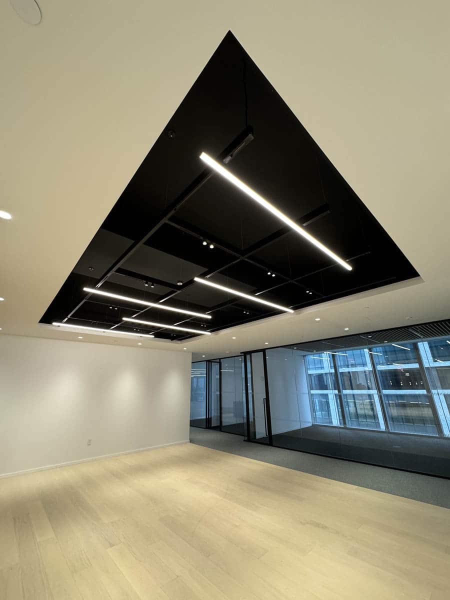A modern, empty office space with light wooden floors, white walls, and a black geometric ceiling feature with linear LED lights. Large glass windows and doors let in natural light, revealing another office building outside.