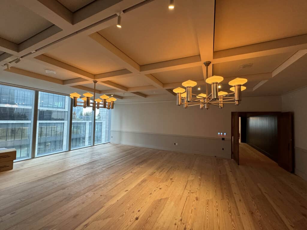 A spacious, empty room with light wood floors, two modern chandeliers, large windows on one side, and warm ceiling lighting, creating a welcoming atmosphere. A doorway leads to another area.