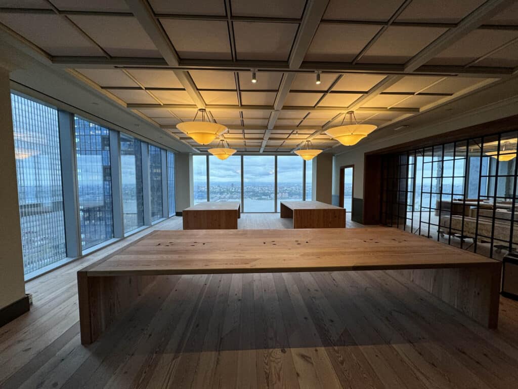 A spacious room with large wooden tables, wood floors, modern ceiling lights, and floor-to-ceiling windows offering a panoramic city view under a cloudy sky.