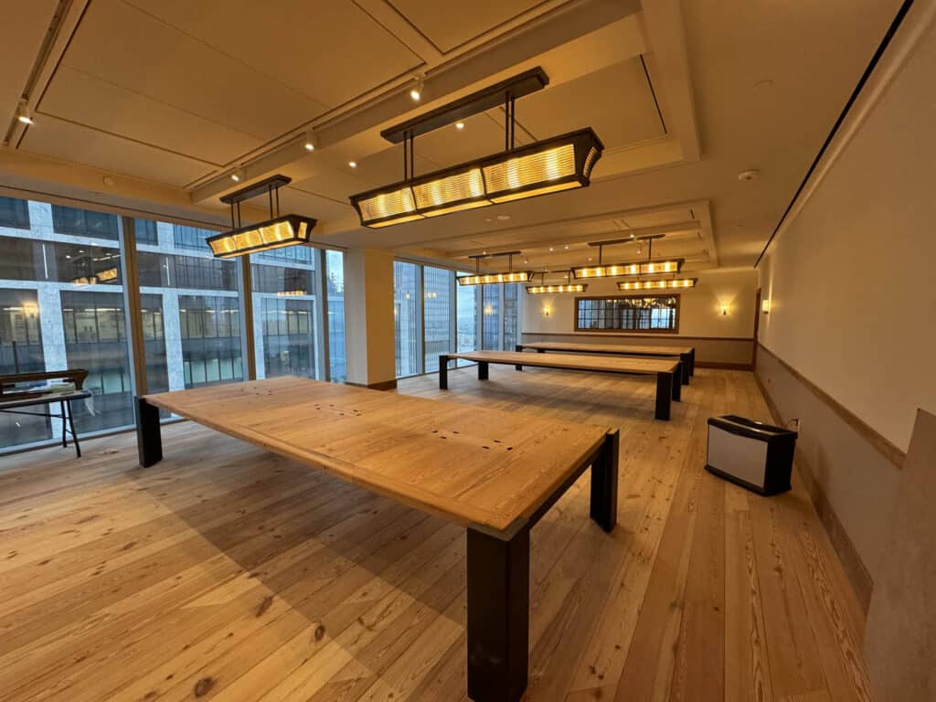 A spacious, well-lit room with large wooden tables, modern rectangular chandeliers, wood flooring, and tall windows providing a view of nearby buildings. The space appears empty and tidy.