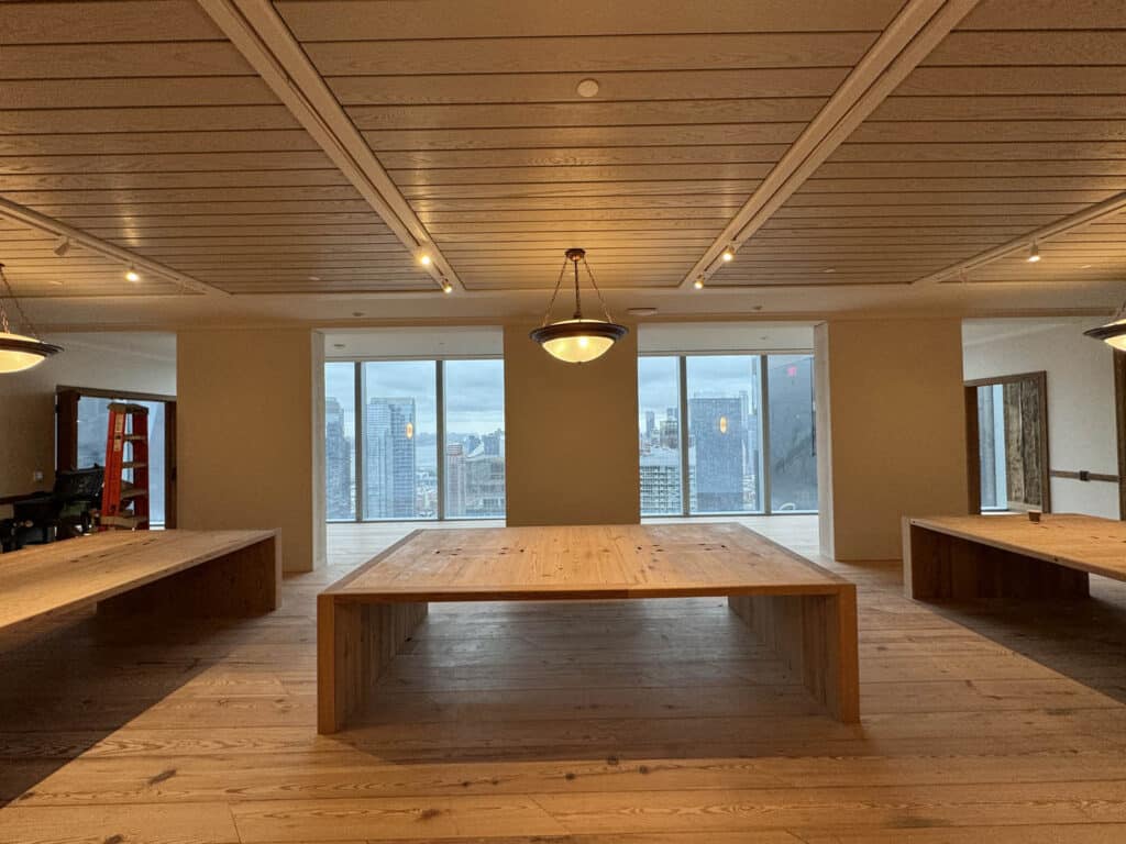 Spacious modern room with large wooden tables, pendant lights, and floor-to-ceiling windows offering a city skyline view. A red ladder and some equipment are visible on the left side.