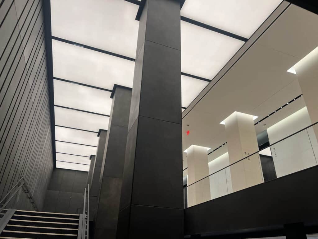 A modern interior with tall, dark columns, a staircase on the left, and a ceiling made of large, illuminated panels. The upper level features glass railings and bright white walls with clean lines.