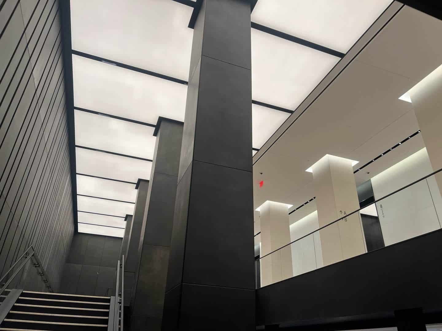 A modern interior with tall, dark columns, a staircase on the left, and a ceiling made of large, illuminated panels. The upper level features glass railings and bright white walls with clean lines.