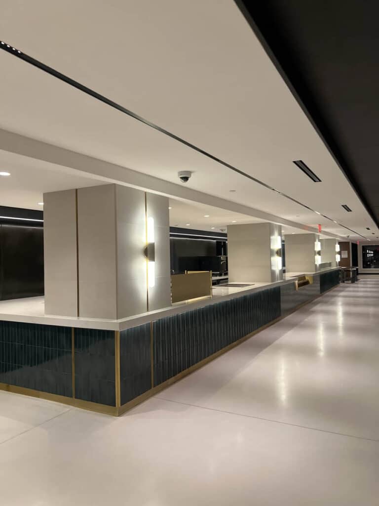 A modern, empty lobby with clean lines, tiled lower walls, beige columns, sleek lighting, and a glossy floor. The space appears minimalistic and well-lit with contemporary decor.