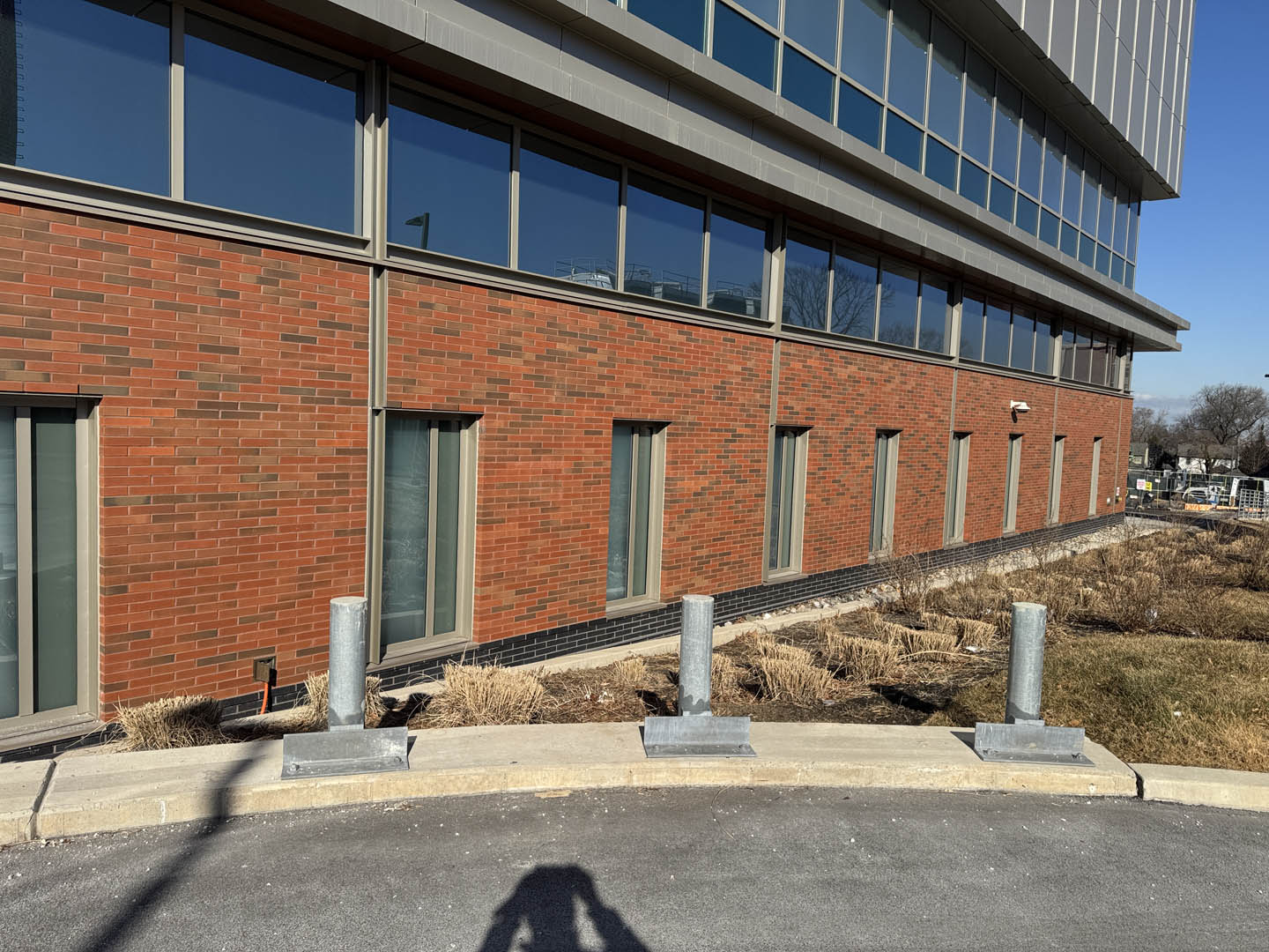A brick building with large windows is bordered by dry grass and small shrubs. Four metal bollards line the edge of a curved driveway, and a shadow of a person taking the photo is visible in the foreground.