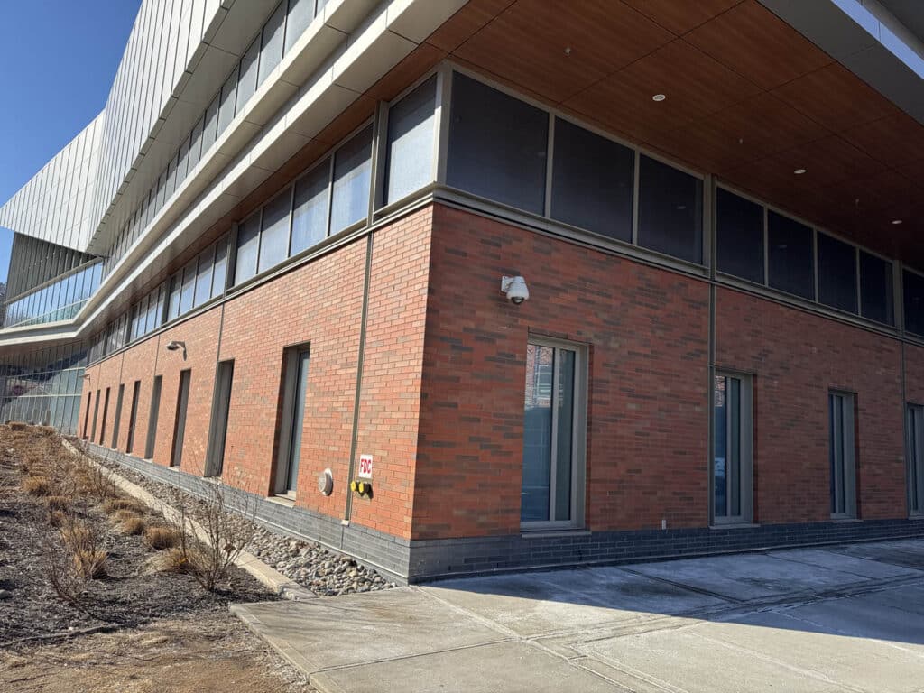 A modern building with red brick and large glass windows on upper floors. The sidewalk and a small landscaped area with sparse plants run alongside the structure under a clear blue sky.