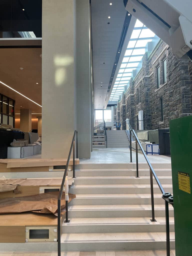 Wide staircase in a modern interior, featuring glass ceiling panels, stone walls on the right, and unfinished construction with boxes, paper, and equipment scattered around. Natural light fills the space.