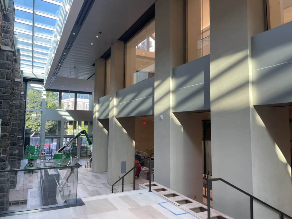 A modern building interior with tall windows, sunlight streaming in, glass walls, stone accents, and workers setting up equipment near the entrance. The space is open, bright, and under construction or renovation.