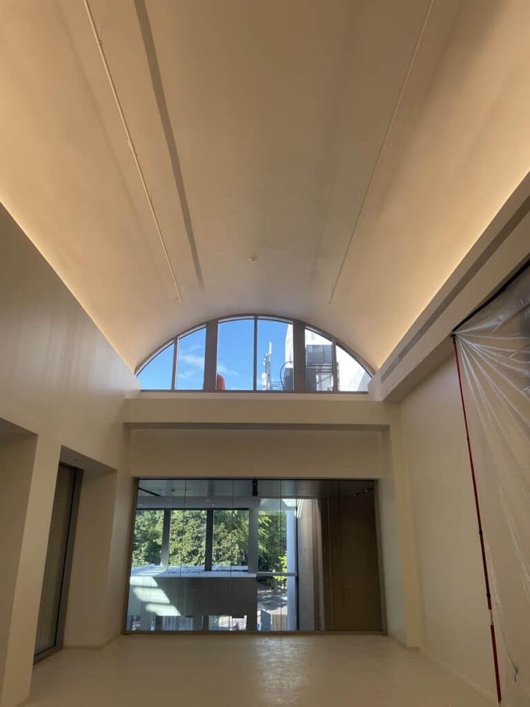 A tall, modern room with a vaulted ceiling, large arched window at the top letting in blue sky, and floor-to-ceiling windows below showing green trees outside. The interior is empty and brightly lit.