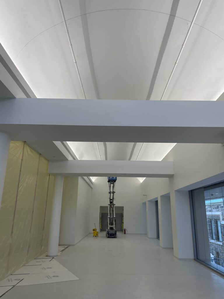 A tall, white, modern interior space with high ceilings and large windows. A scissor lift is positioned in the center, and protective covering is visible on the floor and one wall, indicating ongoing construction or renovation.