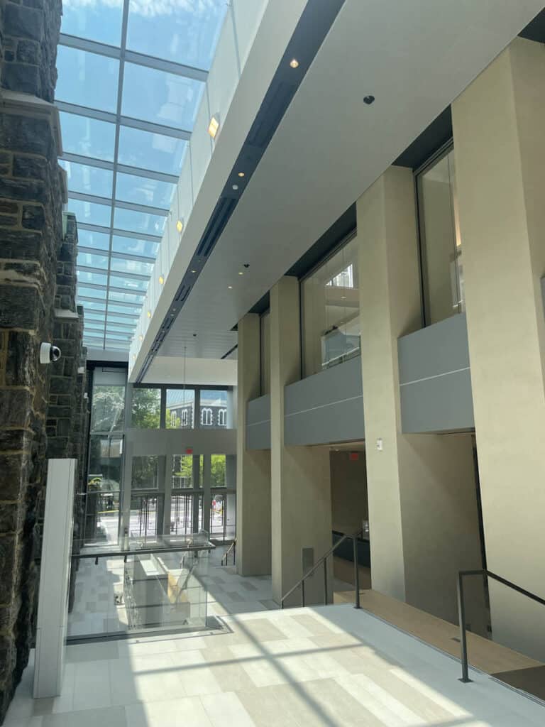 A modern, sunlit building interior with tall windows, glass ceiling, beige walls, a stone wall on the left, and a staircase leading to an outdoor courtyard visible through glass doors.