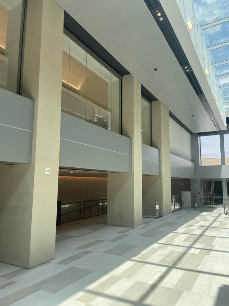 A spacious, modern building interior with tall beige columns, large upper windows, and abundant natural light streaming through a glass ceiling, casting geometric shadows on the light-colored floor.
