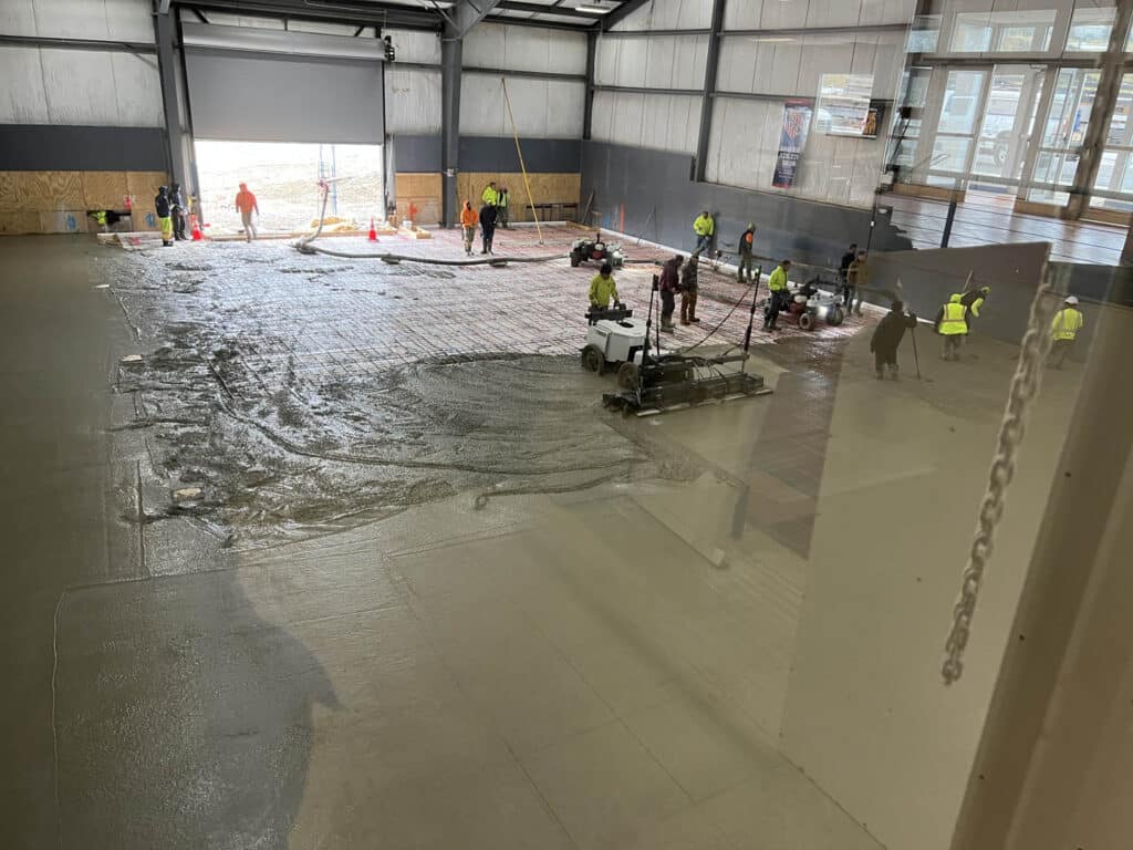 Workers in safety vests pour and smooth concrete inside a large industrial building with high ceilings and open garage doors. Equipment and tools are visible on the wet concrete surface. Natural light enters from outside.