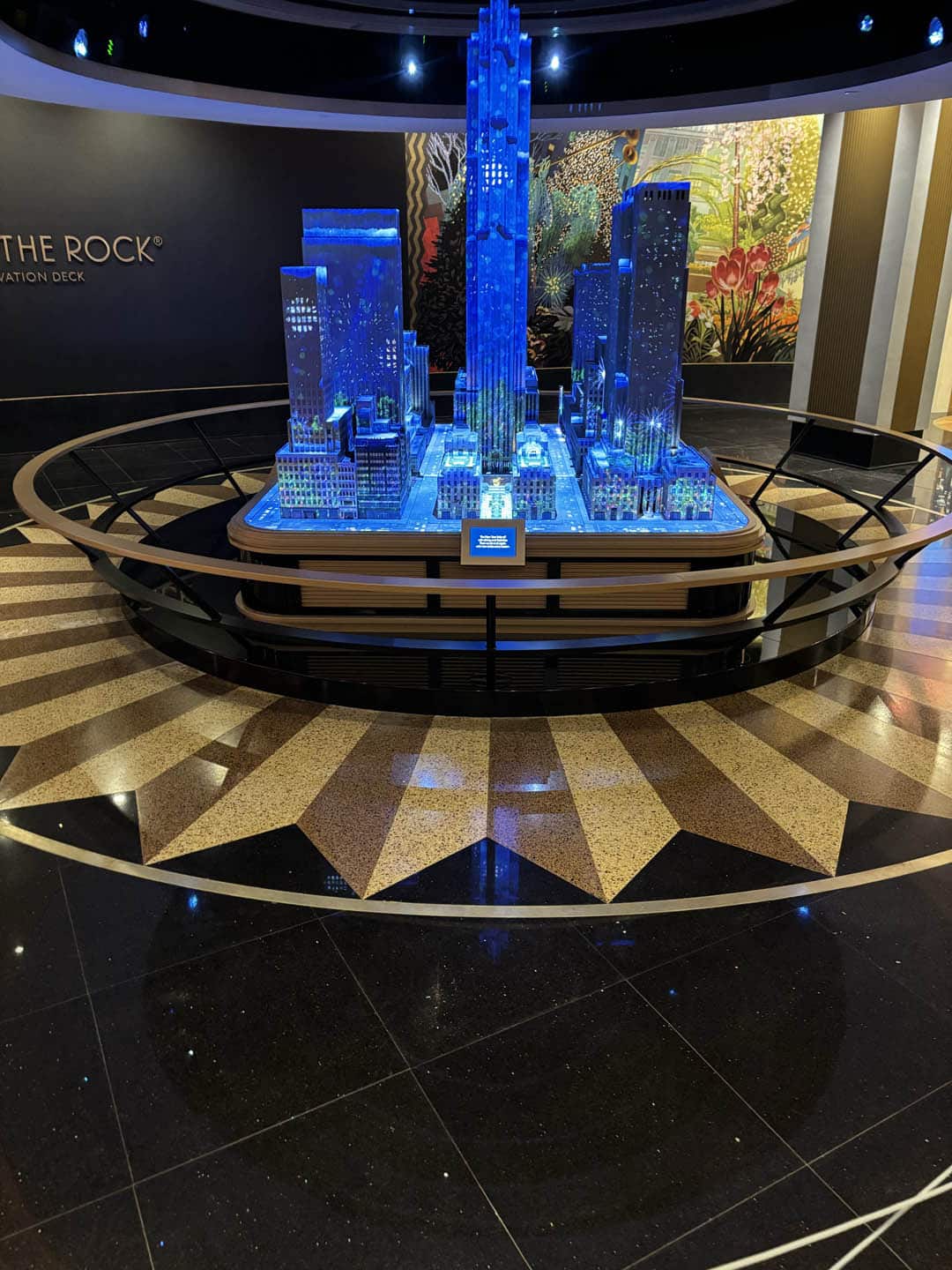A detailed miniature model of a city skyline, illuminated with blue lights, is displayed on a circular platform with a shining starburst pattern on the black and gold floor.