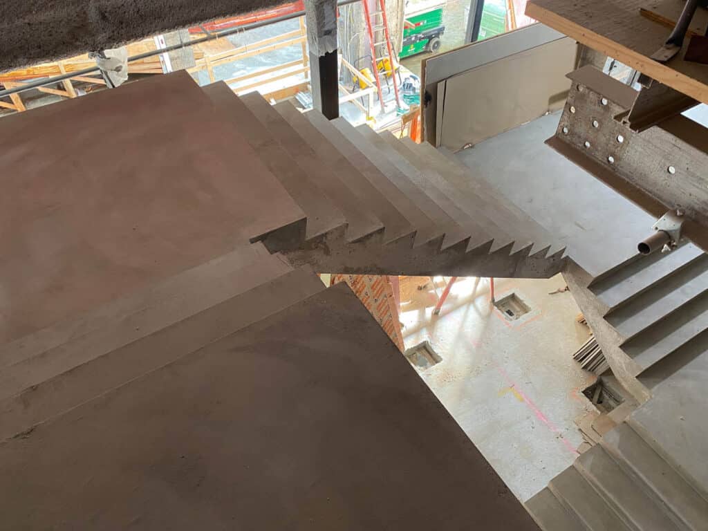 A concrete staircase under construction, with unfinished steps and no railing, connects two floors in a building site. Construction materials and scaffolding are visible in the background.