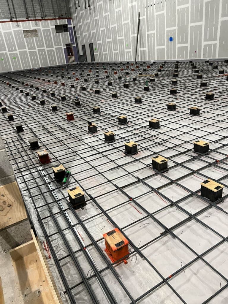 A construction site shows a large grid of steel reinforcement bars on the floor, with small colored boxes placed at grid intersections. The surroundings include unfinished drywall with visible stud lines and markings.