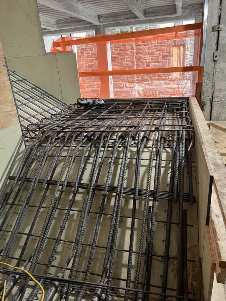 Reinforcement steel bars (rebar) arranged in a grid pattern for a concrete staircase, with temporary wooden and orange safety barriers at a construction site.