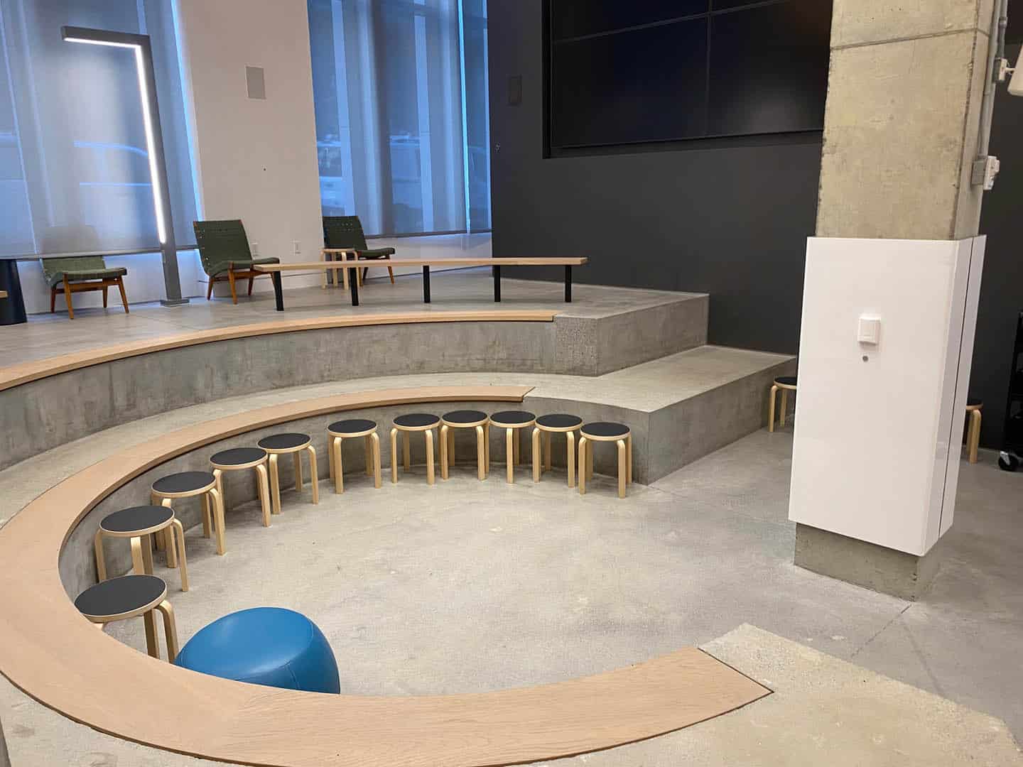 A modern amphitheater-style room with tiered concrete seating, wooden stools arranged in a semi-circle, two green armchairs, large windows, and a concrete pillar with a white electrical box attached.