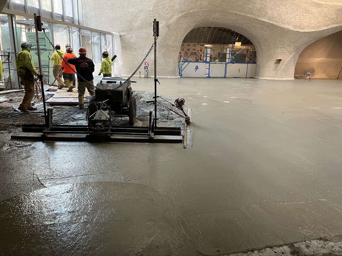 Construction workers smooth freshly poured concrete inside a large, curved architectural space with high ceilings and a large arched opening. Sunlight enters through tall windows on the left side.