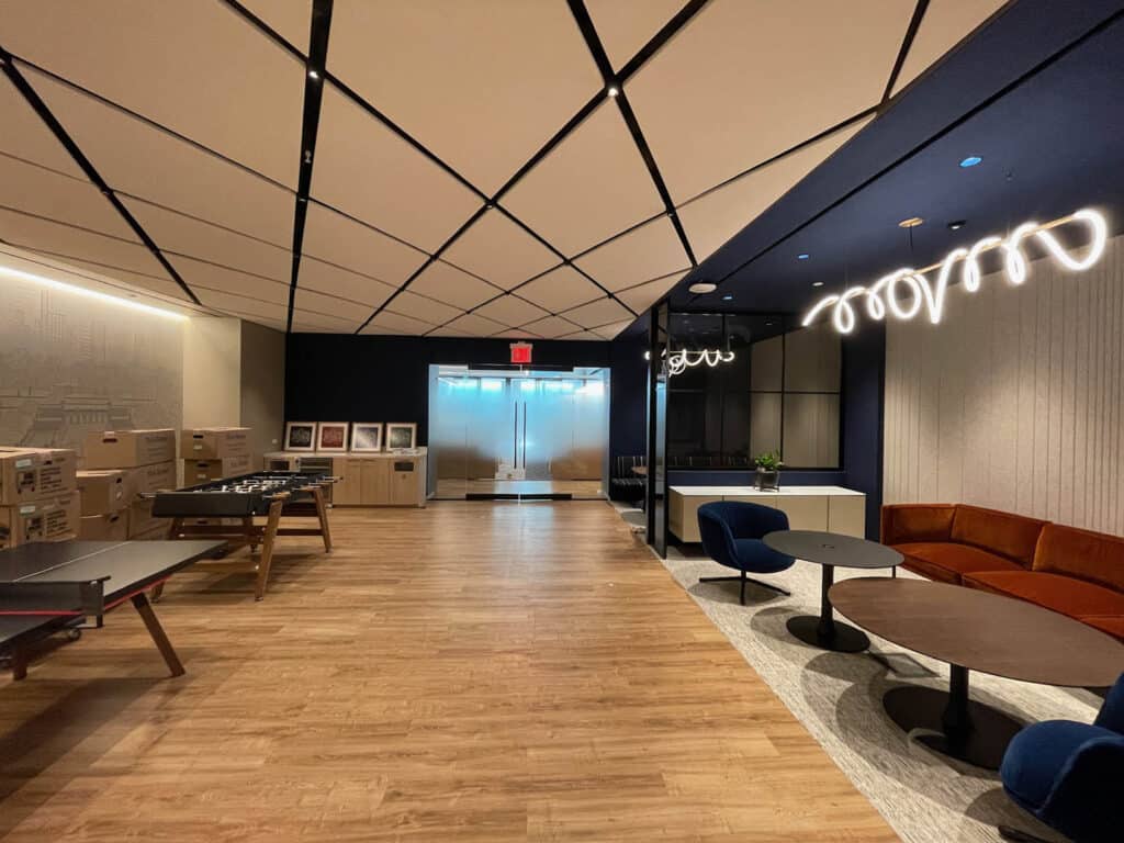 A modern office lounge with wood flooring, a brown couch, round tables, blue chairs, boxes stacked on the left, modern ceiling lights, and double glass doors at the back.