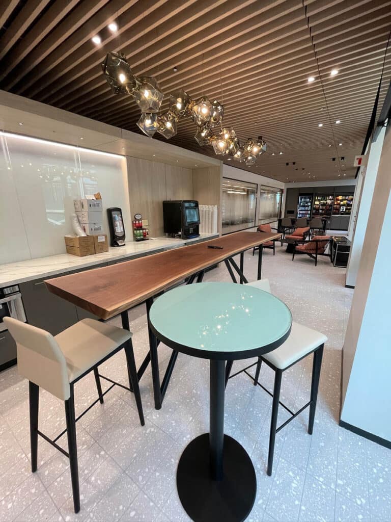 Modern lounge area with a high wooden table, beige barstools, a round glass-top table, unique ceiling light fixtures, drink dispensers along the wall, and seating in the background.