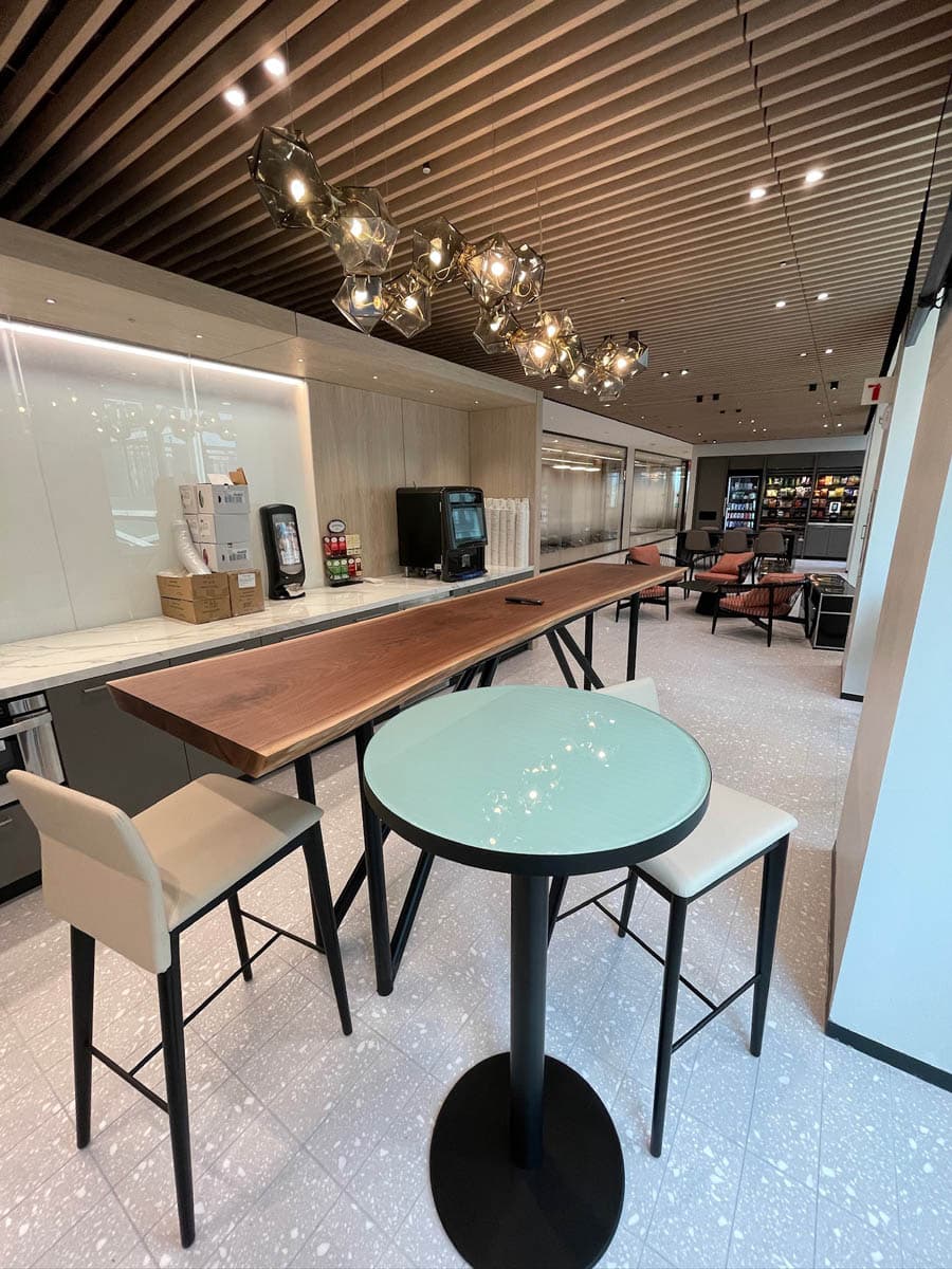 Modern lounge area with a high wooden table, beige barstools, a round glass-top table, unique ceiling light fixtures, drink dispensers along the wall, and seating in the background.