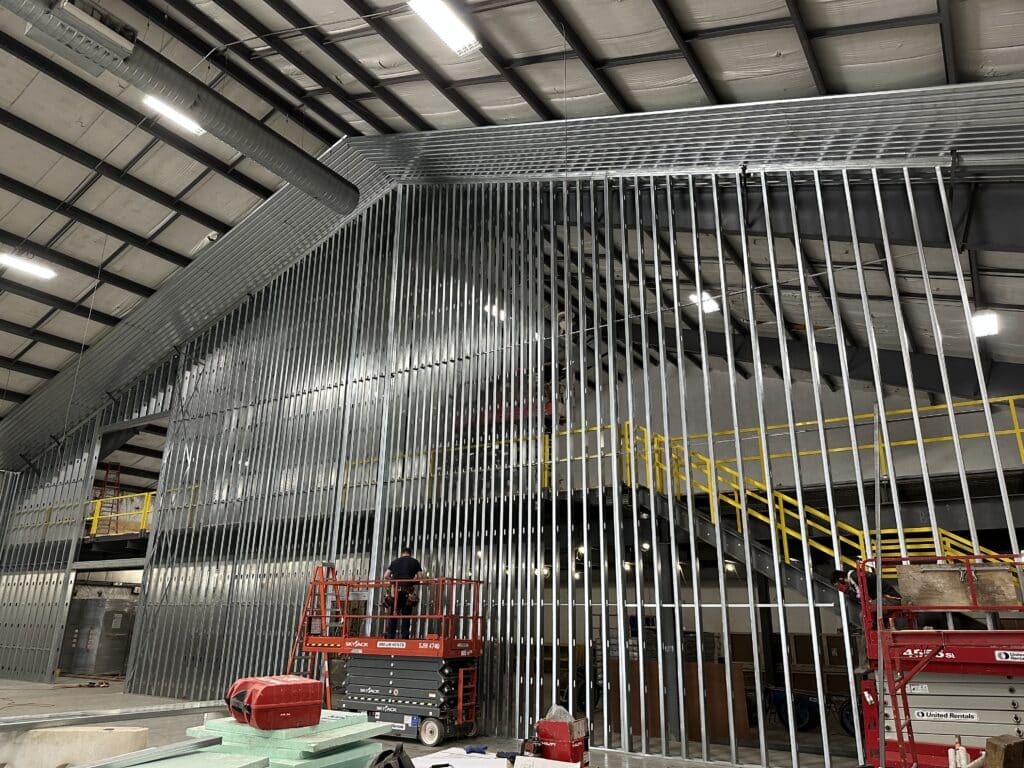 A construction site inside a large building shows tall metal wall framing, yellow railings on an upper level, and workers on red scissor lifts working on the structure. Ceiling beams and ductwork are visible above.