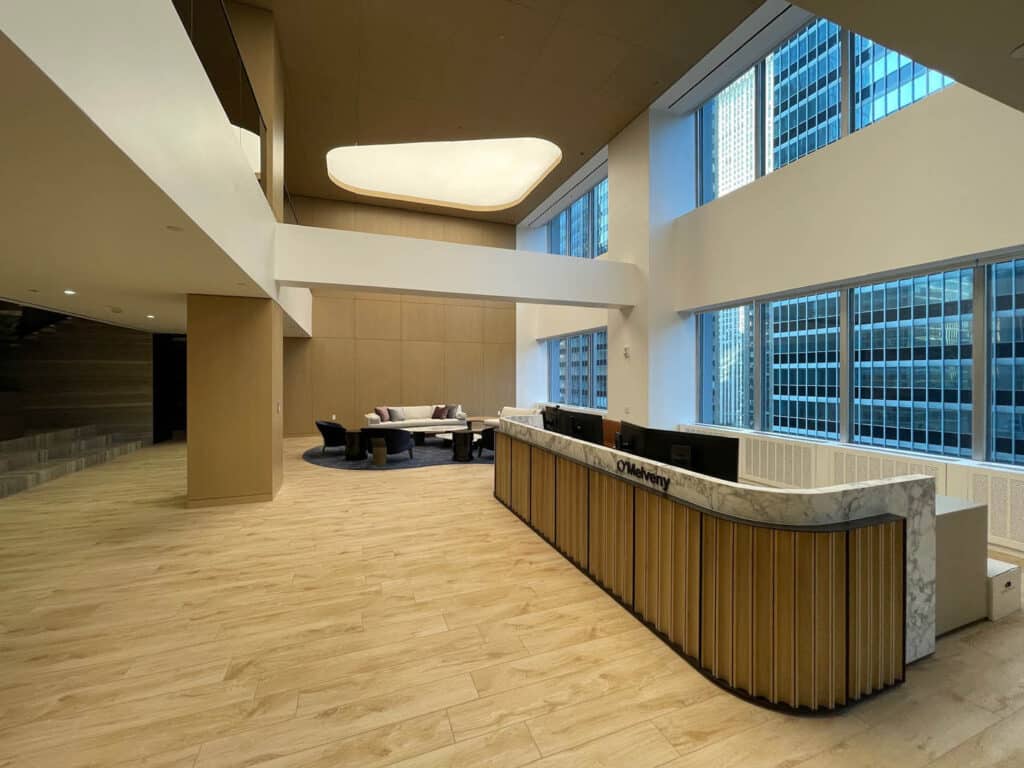 A modern office lobby with a marble reception desk, light wood flooring, tall windows showing city buildings, and a seating area with chairs and a round table in the background.