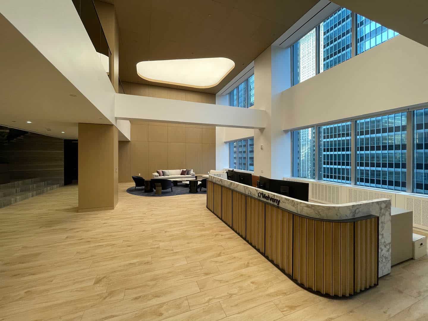 A modern office lobby with a marble reception desk, light wood flooring, tall windows showing city buildings, and a seating area with chairs and a round table in the background.