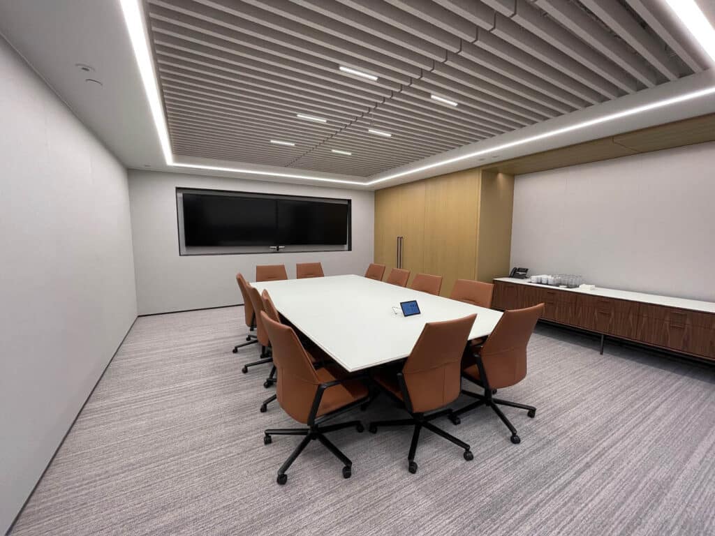 A modern conference room with a long white table surrounded by eight brown office chairs, a large wall-mounted screen, light gray carpet, and minimalist decor. There is a sideboard with refreshments against the far wall.