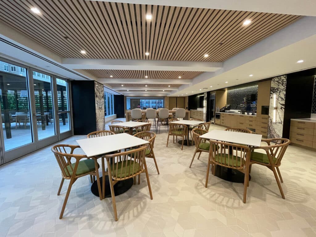 A modern, spacious dining area with wooden tables and chairs, light flooring, and a slatted wood ceiling. Large windows line one side, and a counter with stools and kitchen facilities is visible in the background.