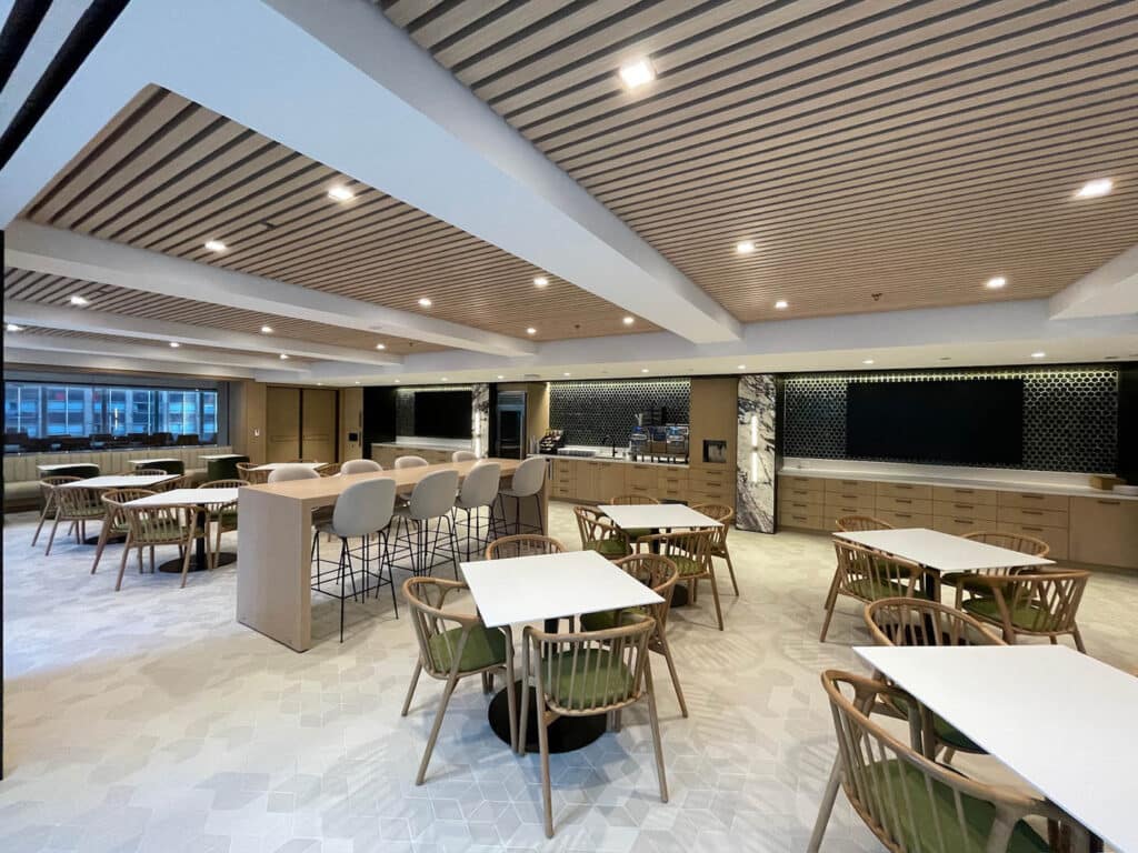 A modern, spacious dining area features wooden tables and chairs, bar seating, and a large, minimalist kitchen along the back wall. Wood paneling on the ceiling and ample lighting create a warm, inviting atmosphere.