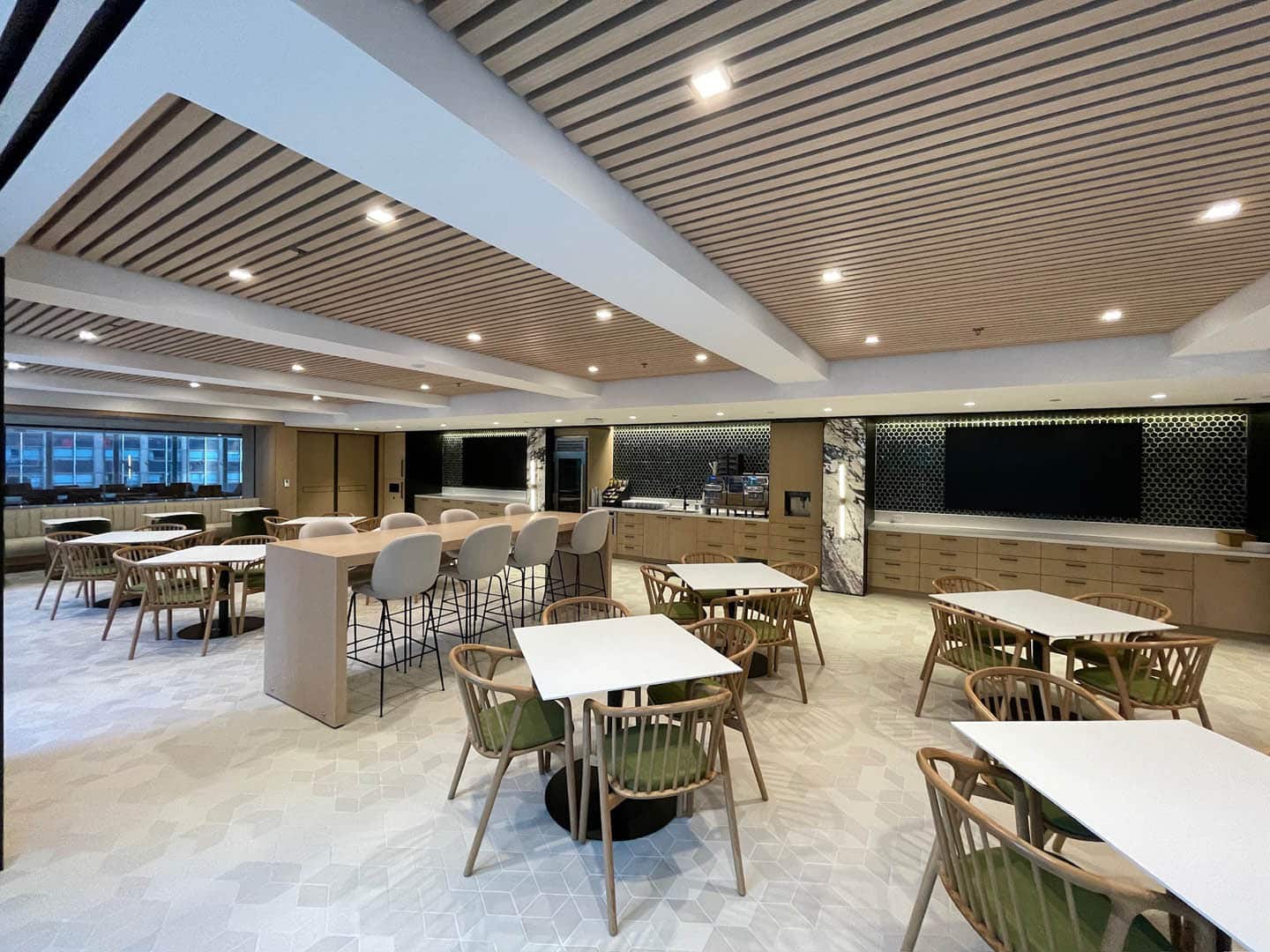 A modern, spacious dining area features wooden tables and chairs, bar seating, and a large, minimalist kitchen along the back wall. Wood paneling on the ceiling and ample lighting create a warm, inviting atmosphere.