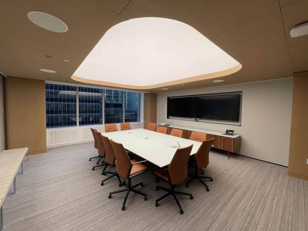A modern conference room with a large white table, ten orange chairs, a wall-mounted TV, and large windows overlooking tall buildings. The room has neutral colors and a distinctive ceiling light.