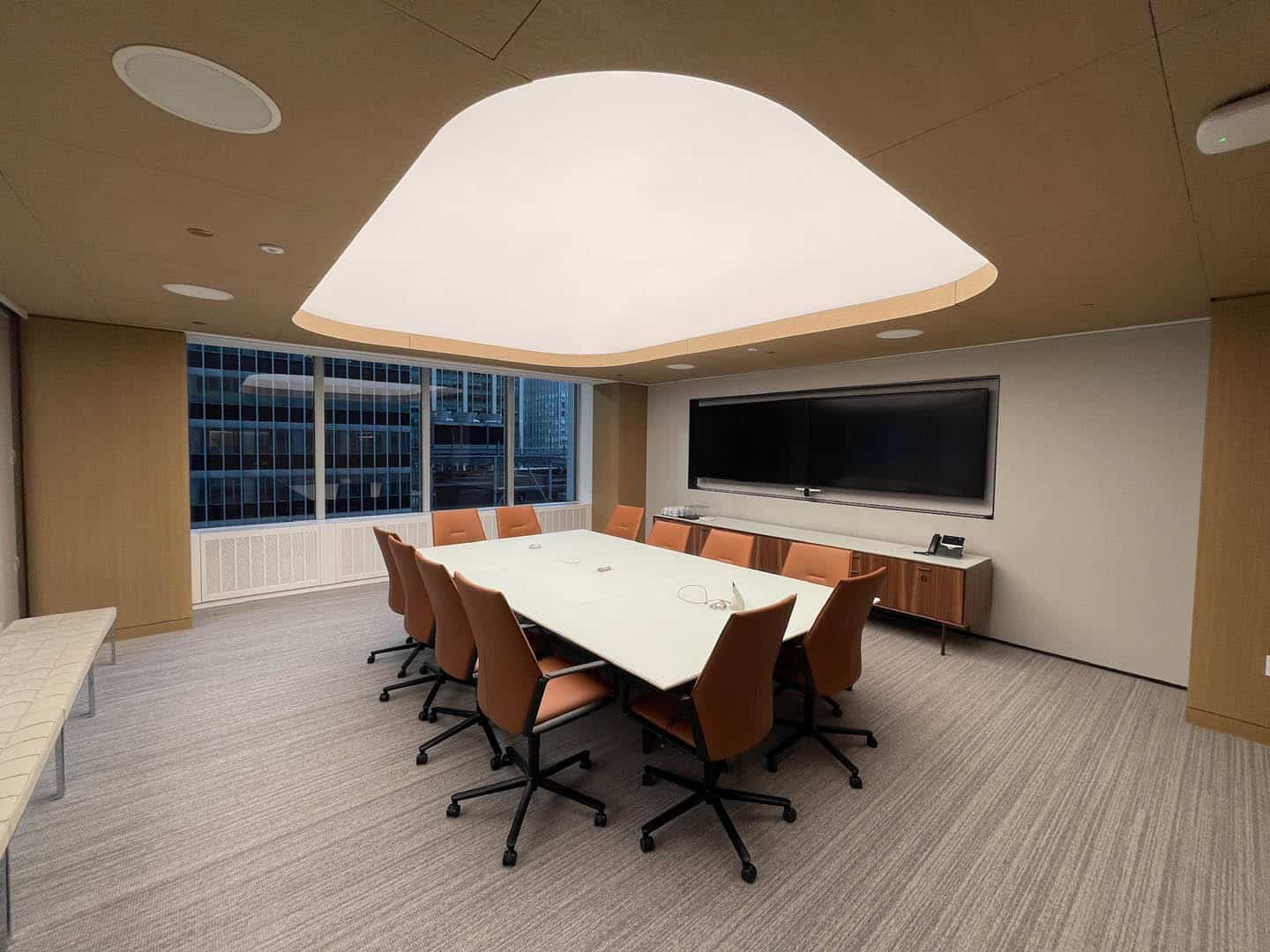 A modern conference room with a large white table, ten orange chairs, a wall-mounted TV, and large windows overlooking tall buildings. The room has neutral colors and a distinctive ceiling light.