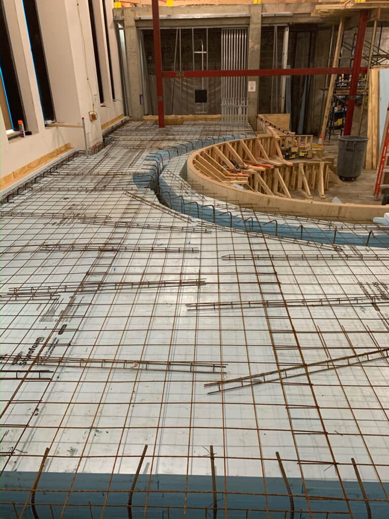 Construction site with a floor covered in blue insulation panels and a grid of steel rebar. Curved wooden forms indicate future concrete pouring areas. The site is indoors, with exposed walls and unfinished sections.