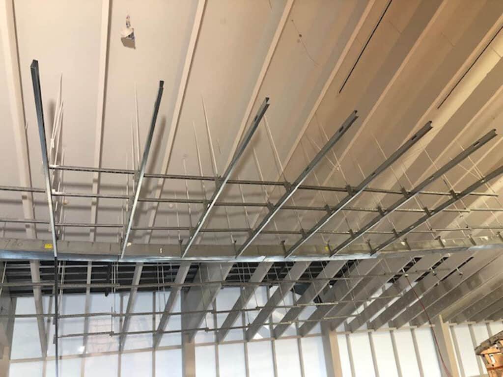 Metal framework for a suspended ceiling is being installed, with grid-like metal bars and rods hanging from a high, unfinished ceiling in a construction site. Natural light comes through large windows in the background.